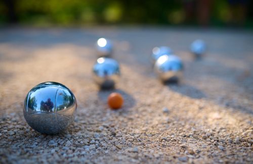 Close-up of petanque balls scattered on gravel with the target ball in focus. The shiny metal balls reflect sunlight, capturing a playful outdoor moment in a park setting.