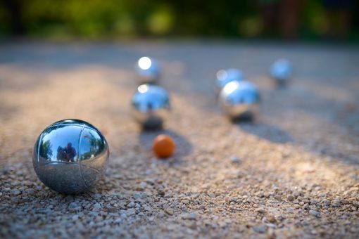 Close-up of petanque balls scattered on gravel with the target ball in focus. The shiny metal balls reflect sunlight, capturing a playful outdoor moment in a park setting.
