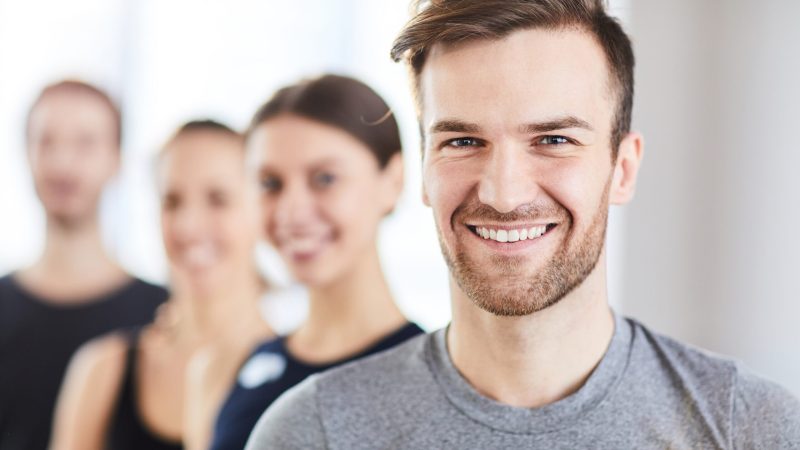 Portrait of cheerful handsome young guy with stubble and fashionable hairstyle smiling at camera while standing in line of fitness trainers